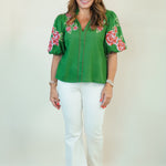 A woman modeling a vibrant green short-sleeve blouse with pink and red floral embroidery on the sleeves and collar, paired with white pants, standing against a plain off-white studio background.