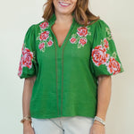 A woman modeling a green short-sleeve button-up blouse with pink and red floral embroidery on the sleeves and collar, paired with white shorts, against a plain off-white studio background.
