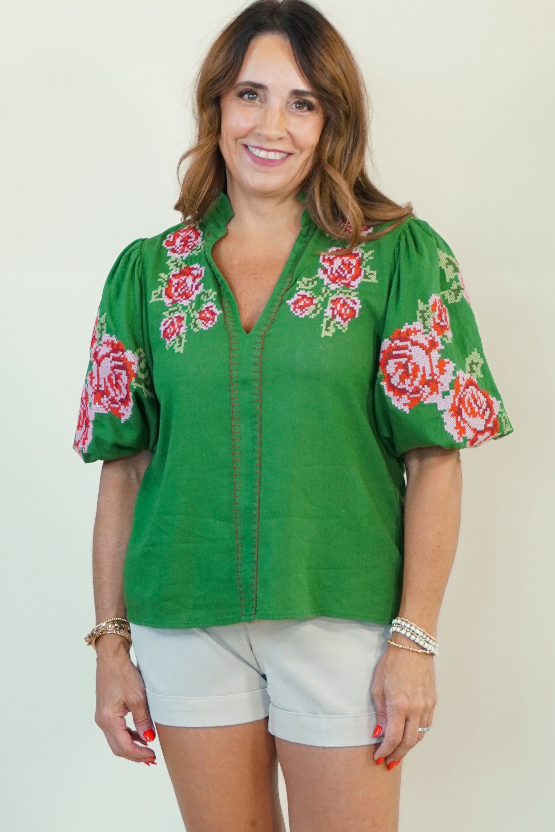 A woman modeling a green short-sleeve button-up blouse with pink and red floral embroidery on the sleeves and collar, paired with white shorts, against a plain off-white studio background.