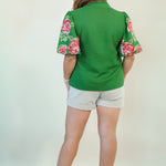 A woman shown from the back wearing a green short-sleeve top with pink and green floral puff sleeves, paired with light gray shorts and brown sandals, against a plain off-white background.