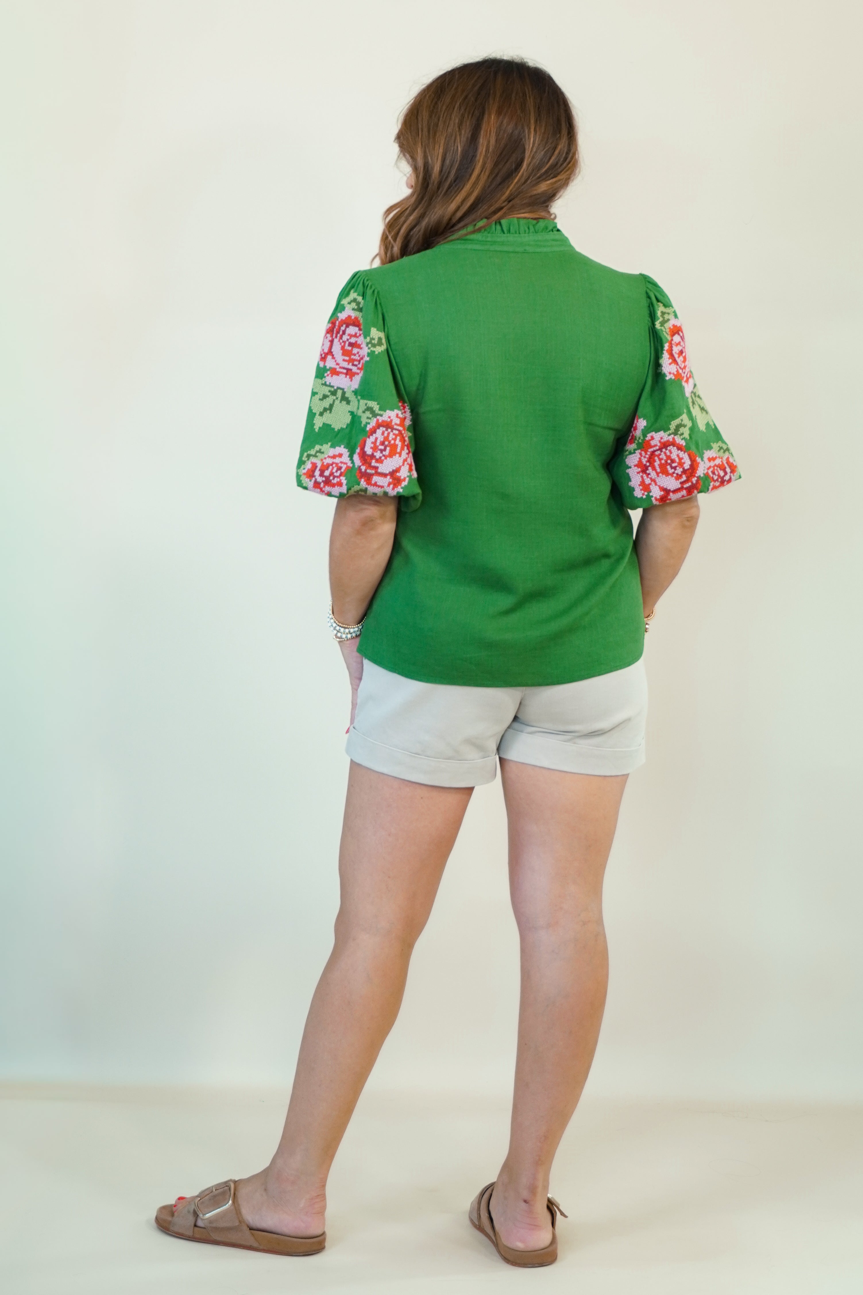 A woman shown from the back wearing a green short-sleeve top with pink and green floral puff sleeves, paired with light gray shorts and brown sandals, against a plain off-white background.