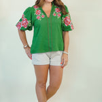 A woman modeling a green short-sleeve blouse with pink and red floral embroidery on the shoulders and sleeves, paired with white shorts, against a plain light beige studio background.