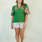 A woman modeling a green short-sleeve blouse with pink and red floral embroidery on the shoulders and sleeves, paired with white shorts and brown sandals, against a plain off-white background.