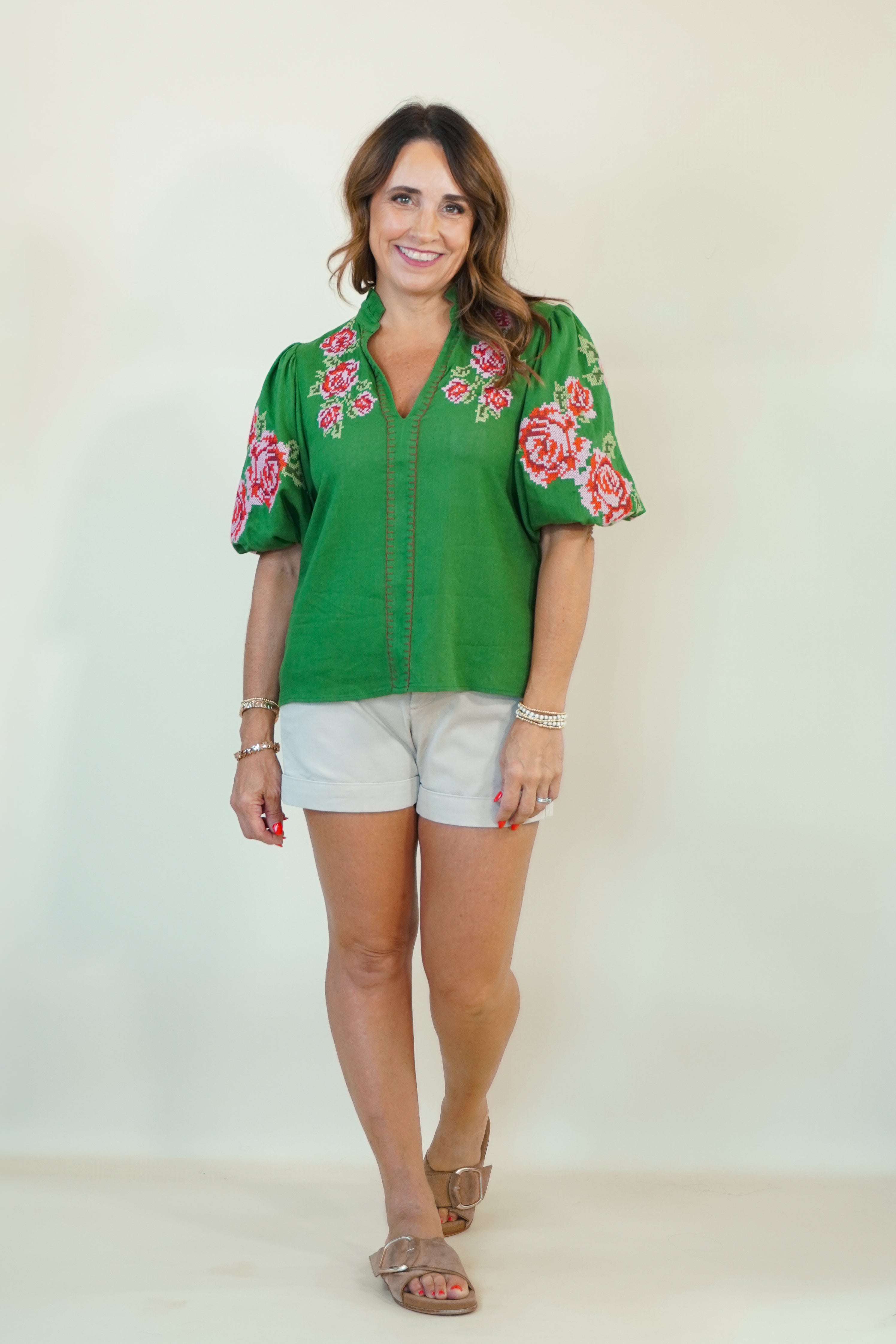A woman modeling a green short-sleeve blouse with pink and red floral embroidery on the shoulders and sleeves, paired with white shorts and brown sandals, against a plain off-white background.