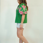 A woman wearing a green short-sleeve blouse with pink and red floral embroidery on the sleeves, paired with white shorts and brown slide sandals, shown in profile against a plain off-white studio background.