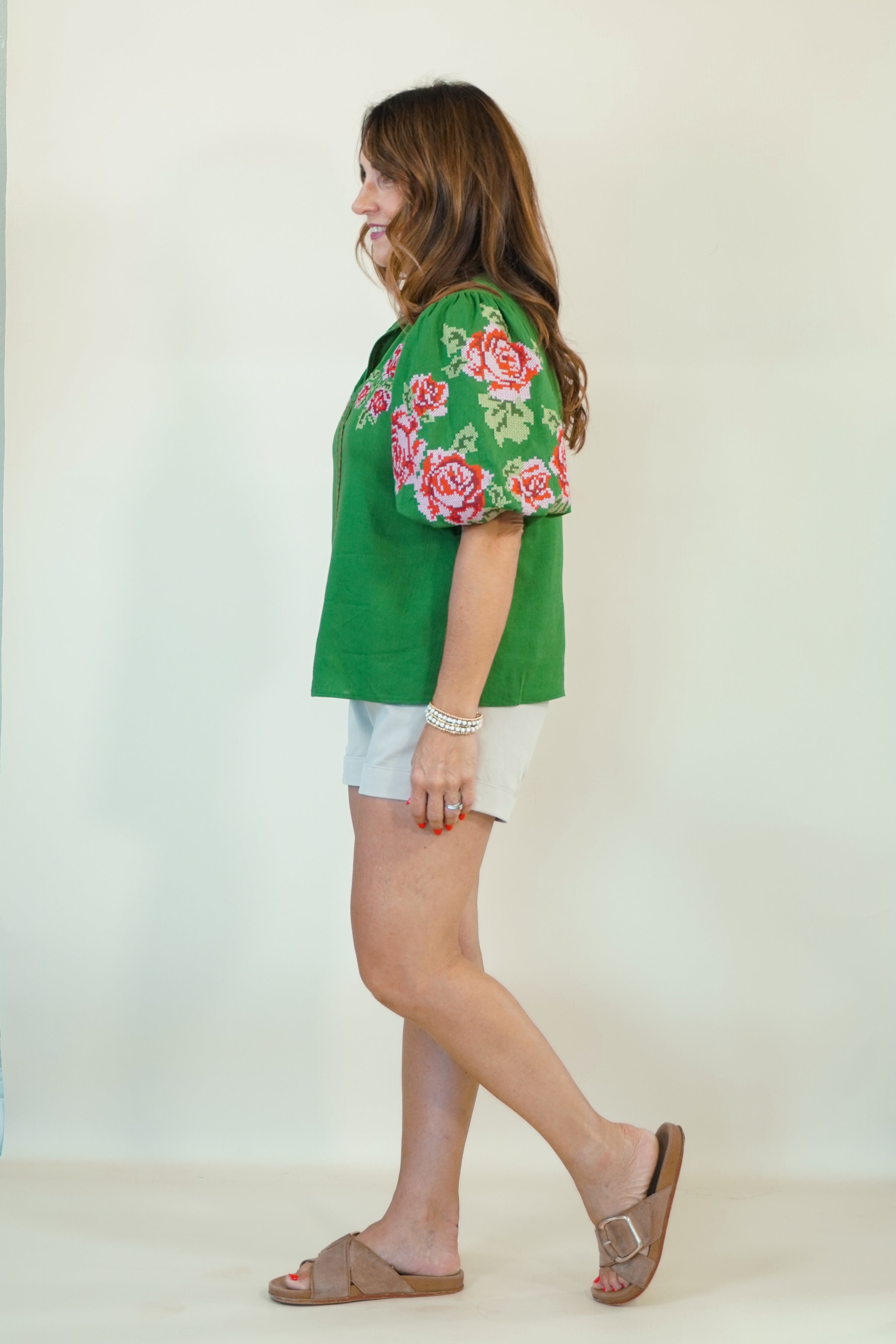 A woman wearing a green short-sleeve blouse with pink and red floral embroidery on the sleeves, paired with white shorts and brown slide sandals, shown in profile against a plain off-white studio background.