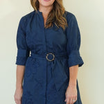 A woman modeling a navy blue midi dress with a matching belt and a subtle tonal floral jacquard pattern, standing against a plain cream-colored background.