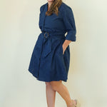 A woman modeling a navy blue wrap-style shirt dress with a matching belt and tie, paired with beige wedge sandals, against a plain off-white studio background.