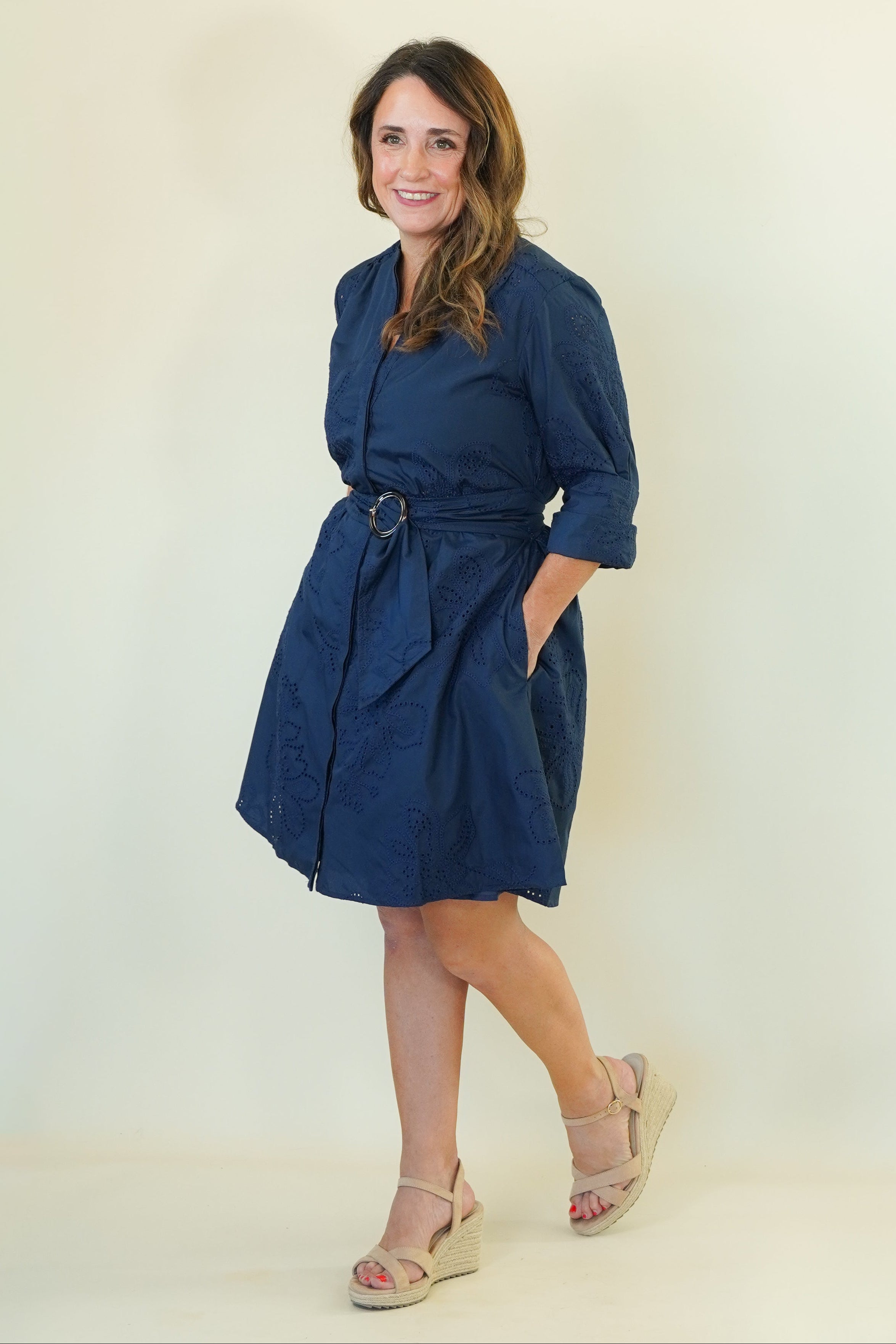 A woman modeling a navy blue wrap-style shirt dress with a matching belt and tie, paired with beige wedge sandals, against a plain off-white studio background.