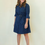 A woman modeling a dark blue belted shirt dress with three-quarter sleeves and a button-up front, paired with beige wedge sandals, standing against a plain cream-colored studio background.