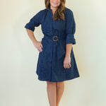 A woman modeling a dark navy blue midi shirt dress with a matching belt and round buckle, standing against a plain cream-colored studio background.