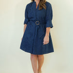 A woman modeling a navy blue short dress with a matching belt and gold-tone ring buckle, standing against a plain off-white studio background.