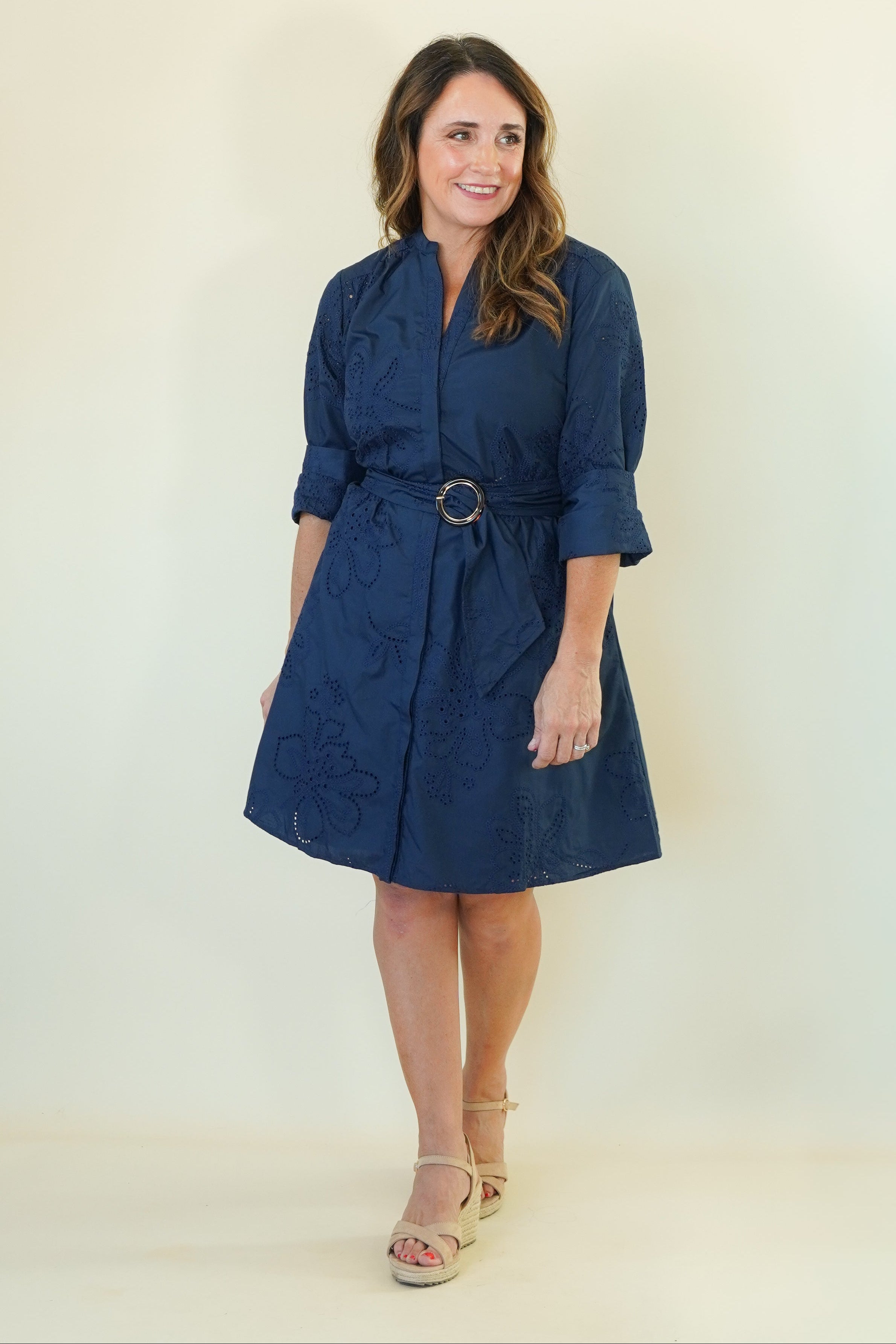 A woman modeling a navy blue short dress with a matching belt and gold-tone ring buckle, standing against a plain off-white studio background.