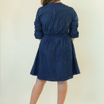 A woman viewed from behind wearing a navy blue long-sleeve dress with a tie waist, paired with beige wedge sandals, against a plain cream background.