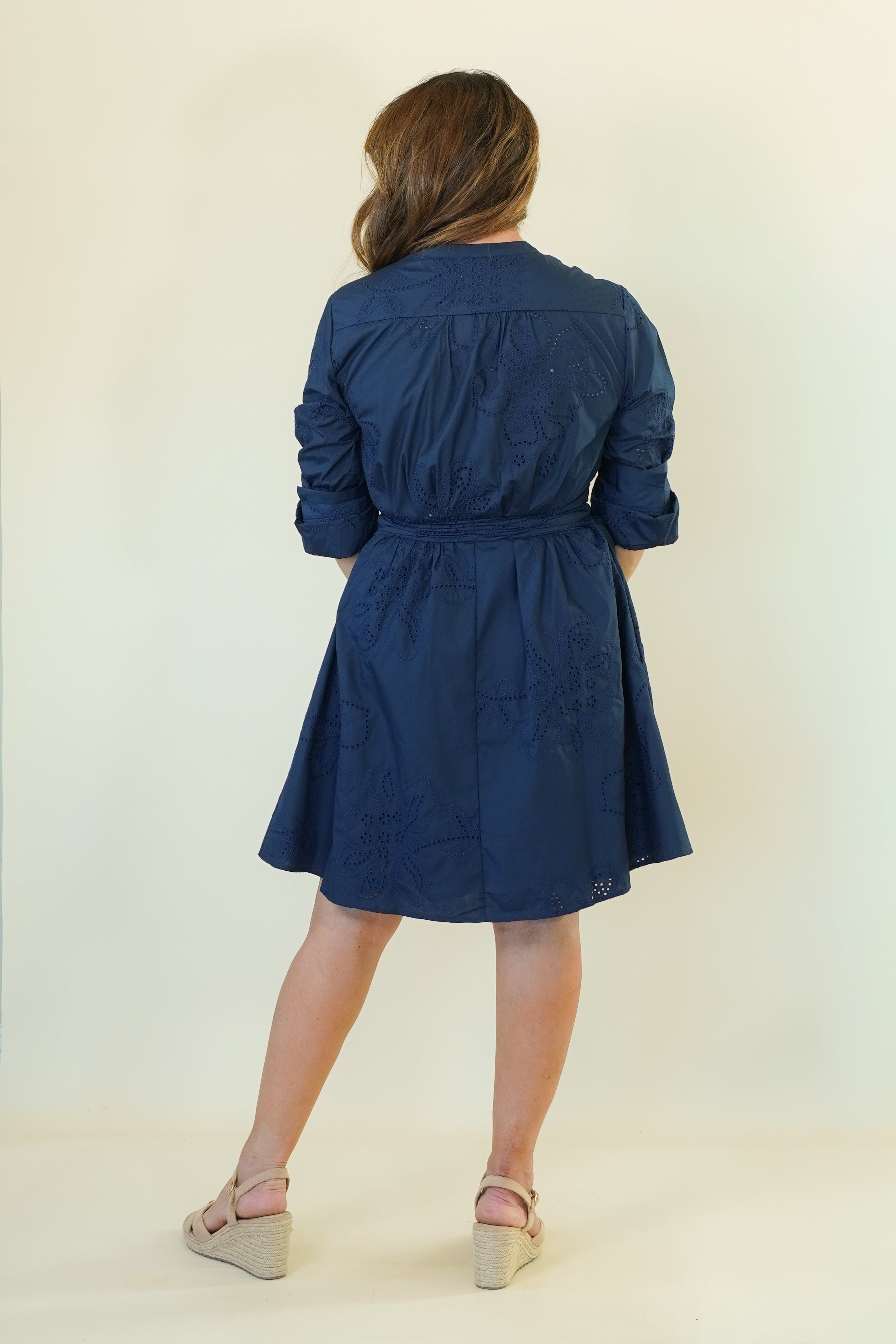 A woman viewed from behind wearing a navy blue long-sleeve dress with a tie waist, paired with beige wedge sandals, against a plain cream background.