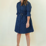 A woman modeling a navy blue belted dress with three-quarter sleeves and a shawl collar, paired with beige wedge sandals, against a plain cream studio background.