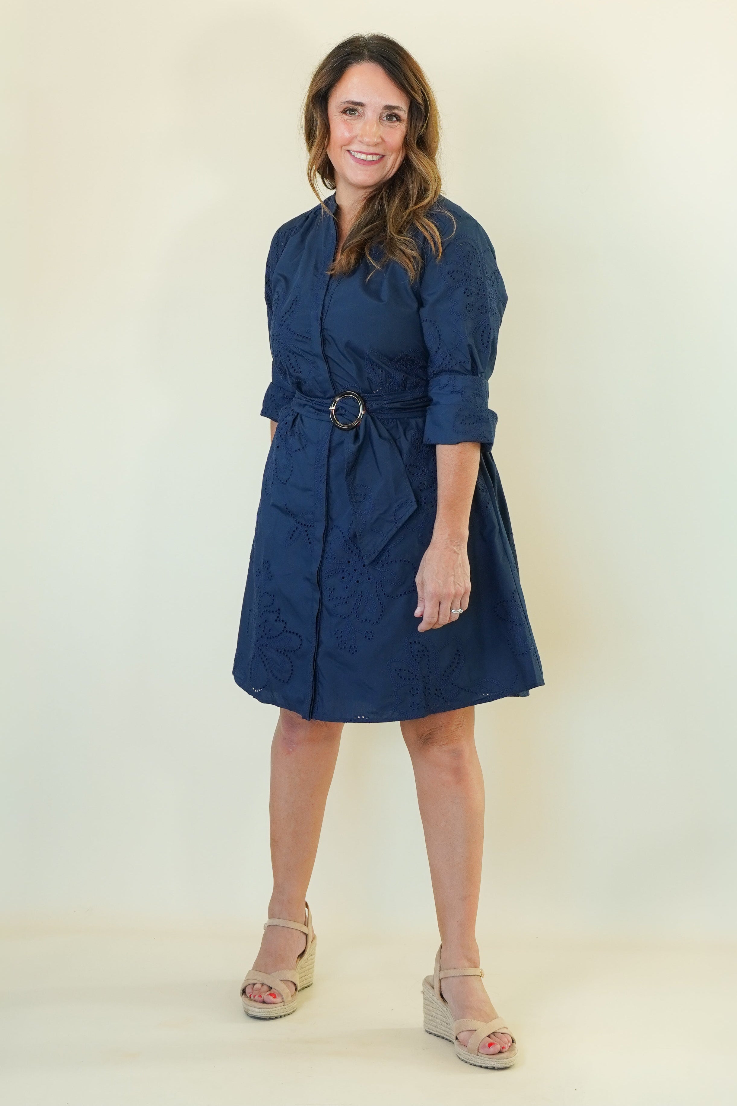 A woman modeling a navy blue belted dress with three-quarter sleeves and a shawl collar, paired with beige wedge sandals, against a plain cream studio background.