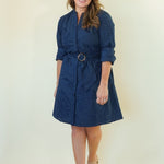 A woman modeling a dark navy blue textured dress with a matching belt and round buckle, standing against a plain cream-colored background.