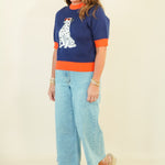 A woman modeling a navy blue knit sweater with orange trim and a white Dalmatian graphic, paired with light blue cropped jeans and beige wedge sandals, against a plain cream background.