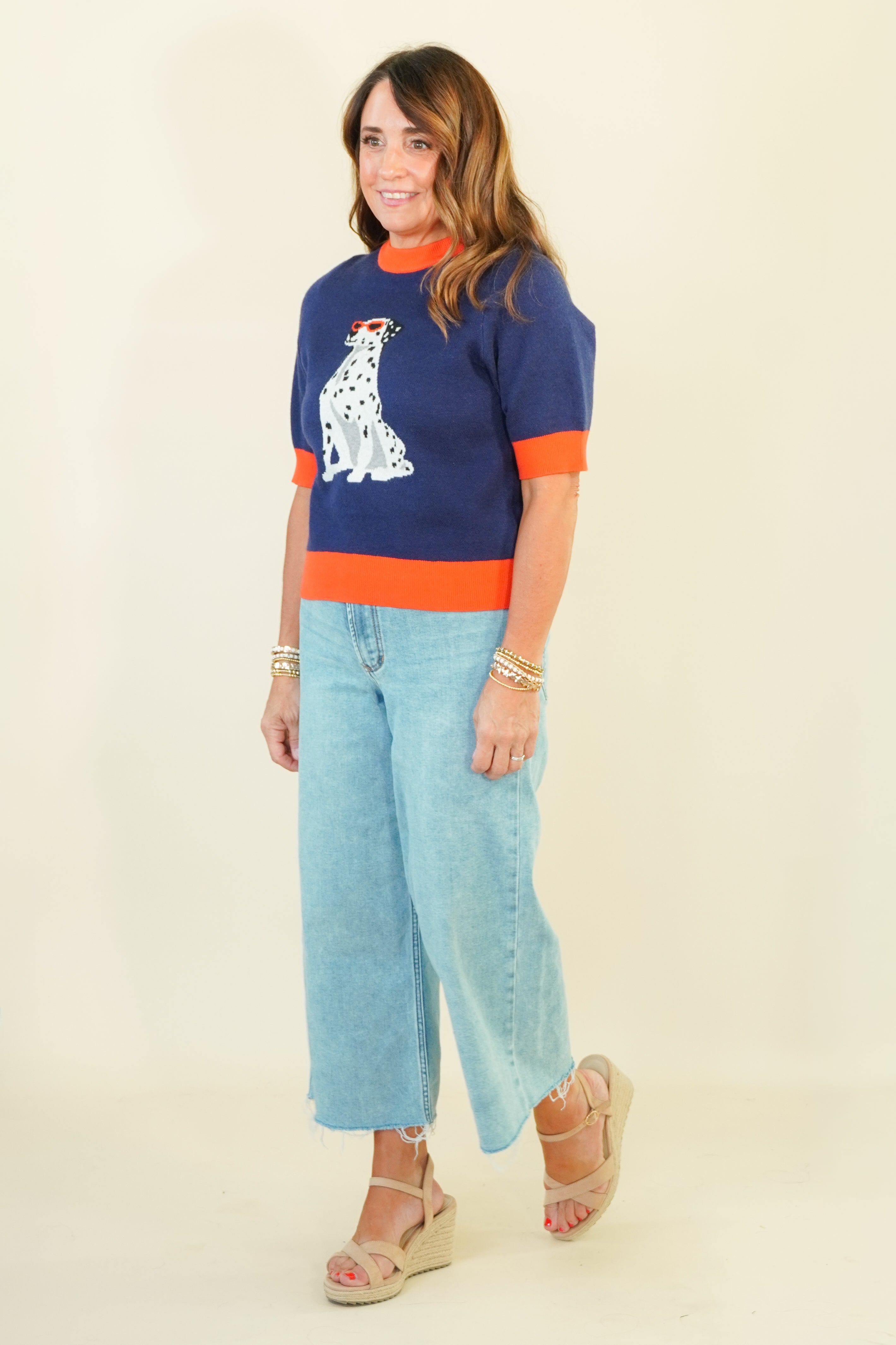 A woman modeling a navy blue knit sweater with orange trim and a white Dalmatian graphic, paired with light blue cropped jeans and beige wedge sandals, against a plain cream background.