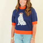 A woman modeling a navy blue short-sleeve knit sweater with orange trim and a white Dalmatian graphic on the front, paired with light blue wide-leg jeans and beige sandals, against a plain cream background.