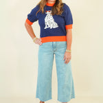 A woman modeling a navy blue short-sleeve knit sweater with orange trim and a white Dalmatian dog graphic, paired with light blue wide-leg jeans and beige wedge sandals, against a plain cream background.