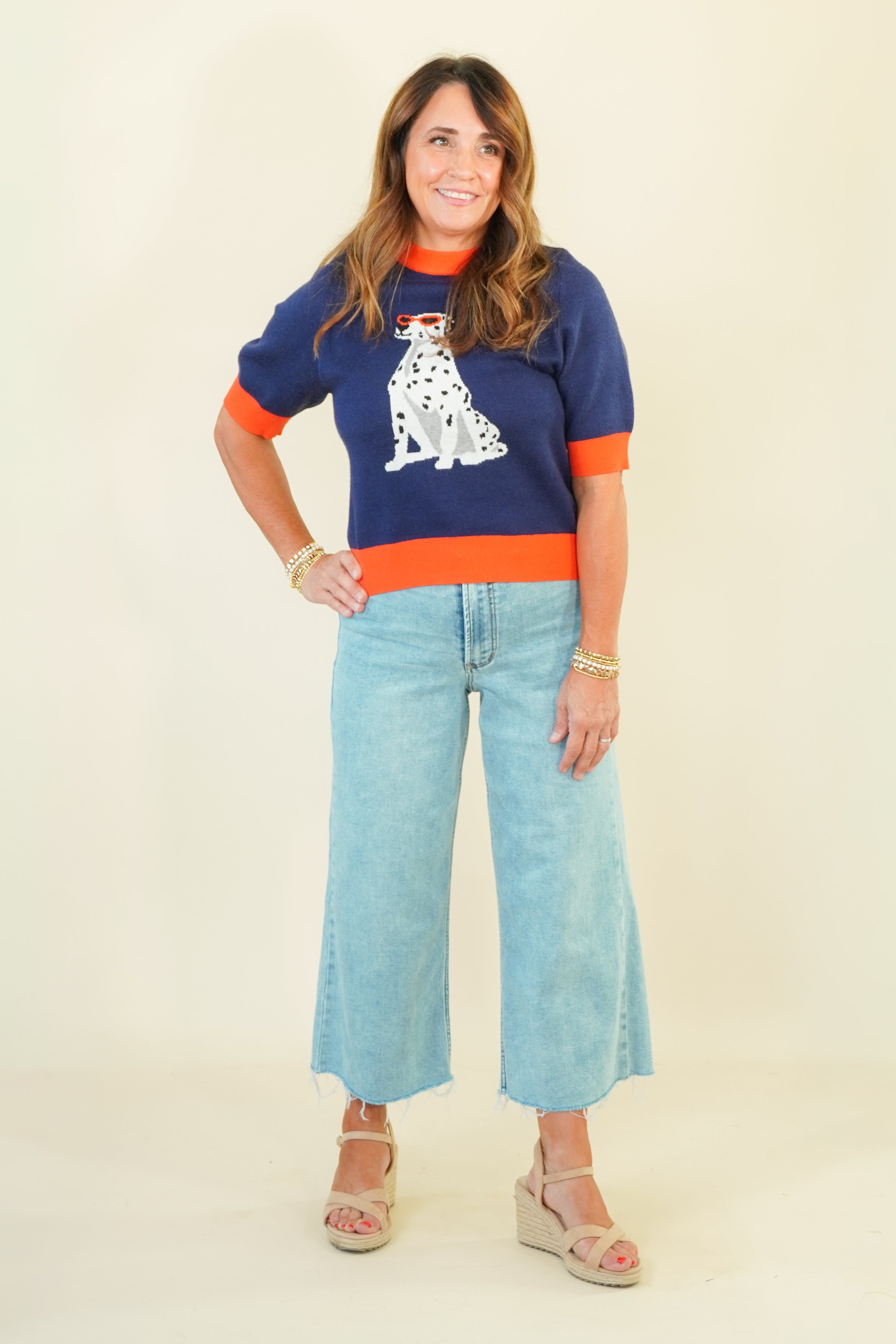 A woman modeling a navy blue short-sleeve knit sweater with orange trim and a white Dalmatian dog graphic, paired with light blue wide-leg jeans and beige wedge sandals, against a plain cream background.