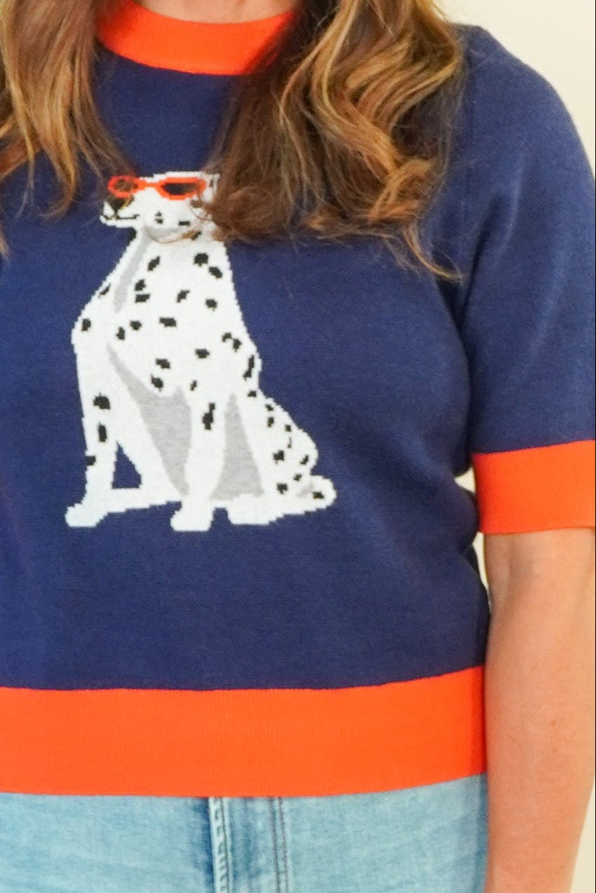 A woman modeling a navy blue knit sweater with orange trim and a white Dalmatian graphic, paired with light blue wide-leg jeans and beige sandals, against a plain cream background.
