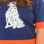 A woman modeling a navy blue knit sweater with orange trim and a white Dalmatian graphic, paired with light blue wide-leg jeans and beige sandals, against a plain cream background.