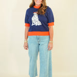 A woman modeling a navy blue knit sweater with orange trim and a white Dalmatian graphic, paired with light blue cropped jeans and beige wedge sandals, against a plain cream background.