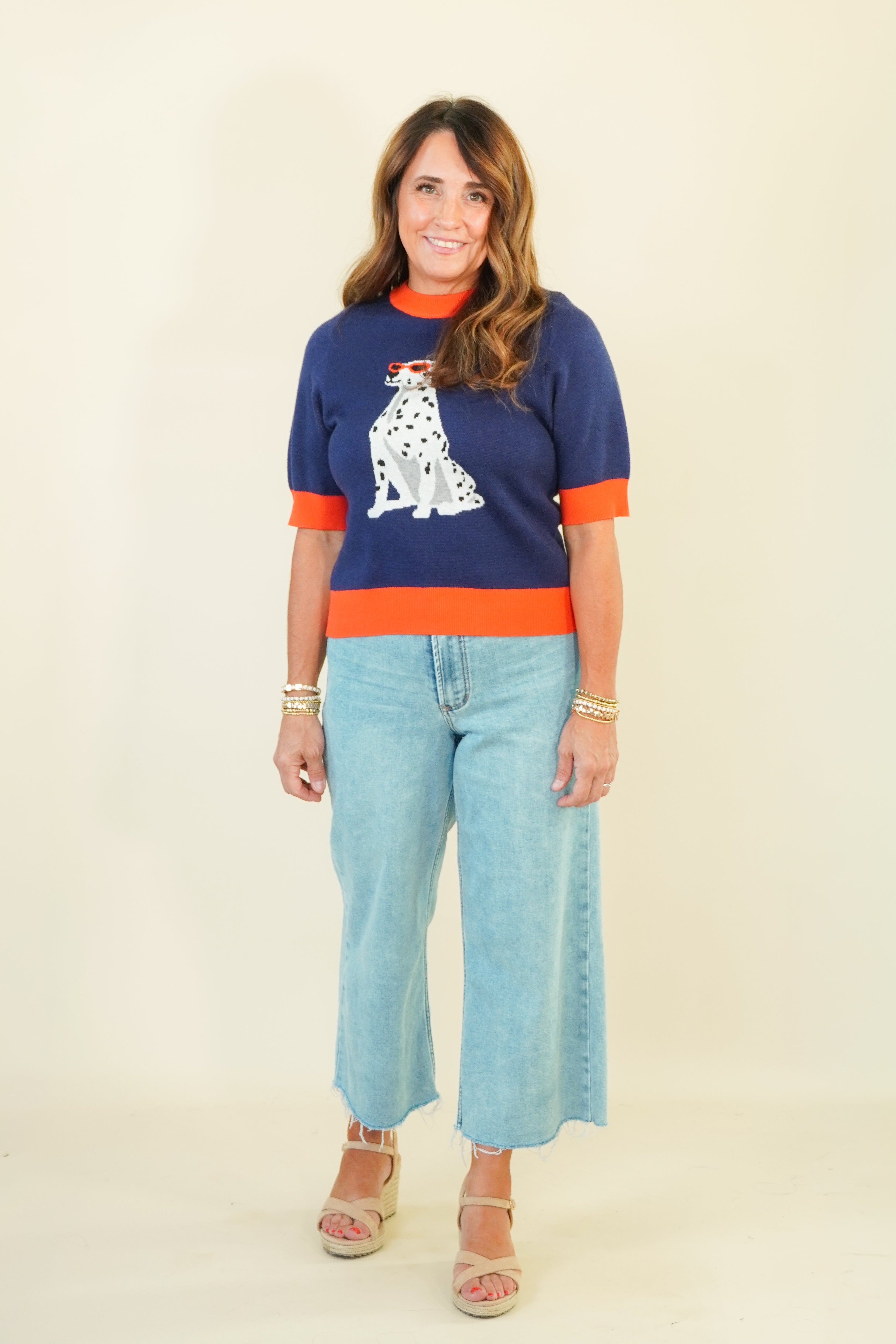 A woman modeling a navy blue knit sweater with orange trim and a white Dalmatian graphic, paired with light blue cropped jeans and beige wedge sandals, against a plain cream background.