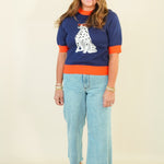 A woman modeling a navy blue short-sleeve knit sweater with bright orange trim and a white Dalmatian graphic on the front, paired with light blue cropped jeans and beige sandals, against a plain cream background.