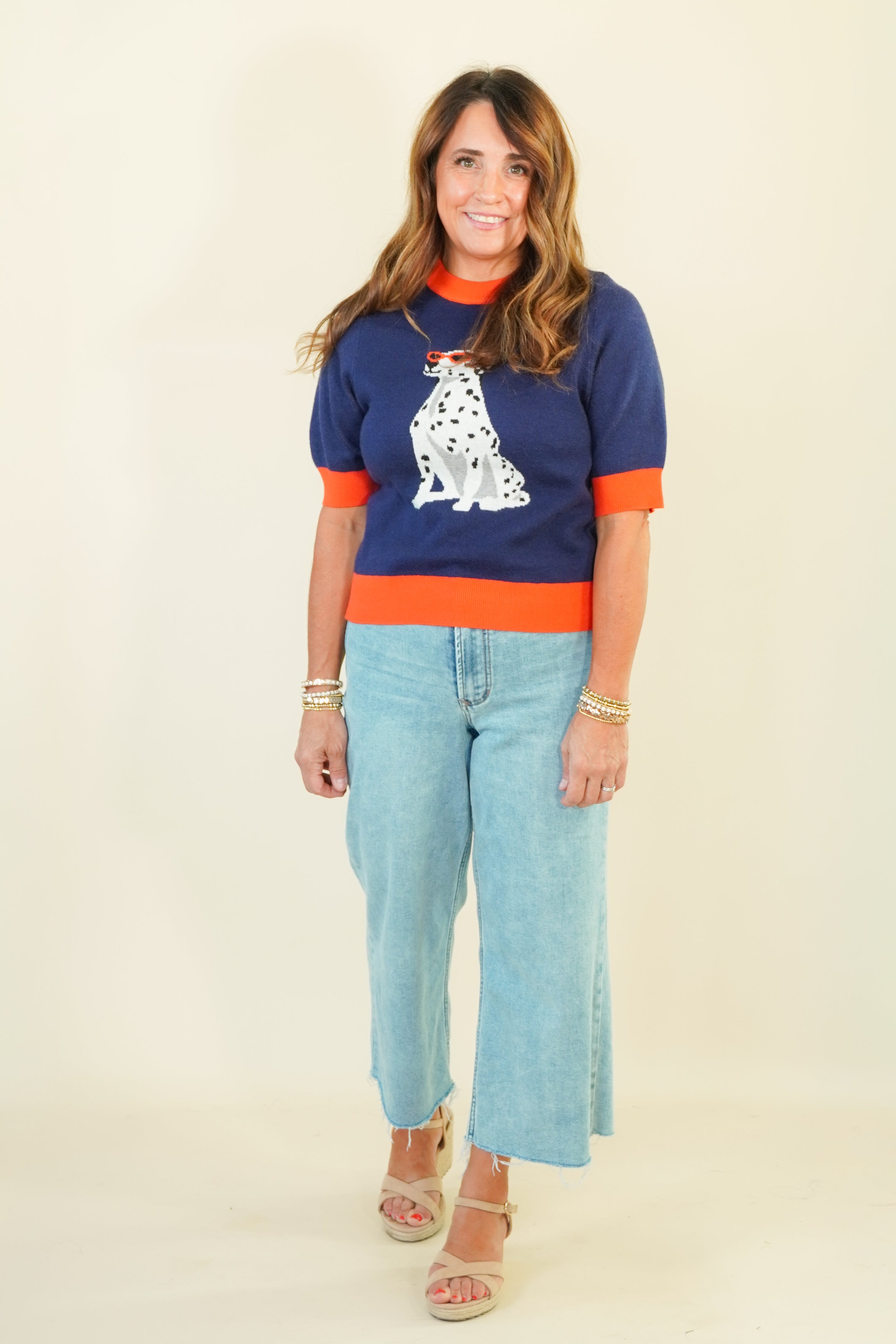 A woman modeling a navy blue short-sleeve knit sweater with bright orange trim and a white Dalmatian graphic on the front, paired with light blue cropped jeans and beige sandals, against a plain cream background.