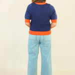 A woman viewed from behind wearing a short-sleeve knit sweater with a navy blue body and bright orange trim at the collar, cuffs, and hem, paired with light blue cropped jeans and beige wedge sandals, against a plain cream background.