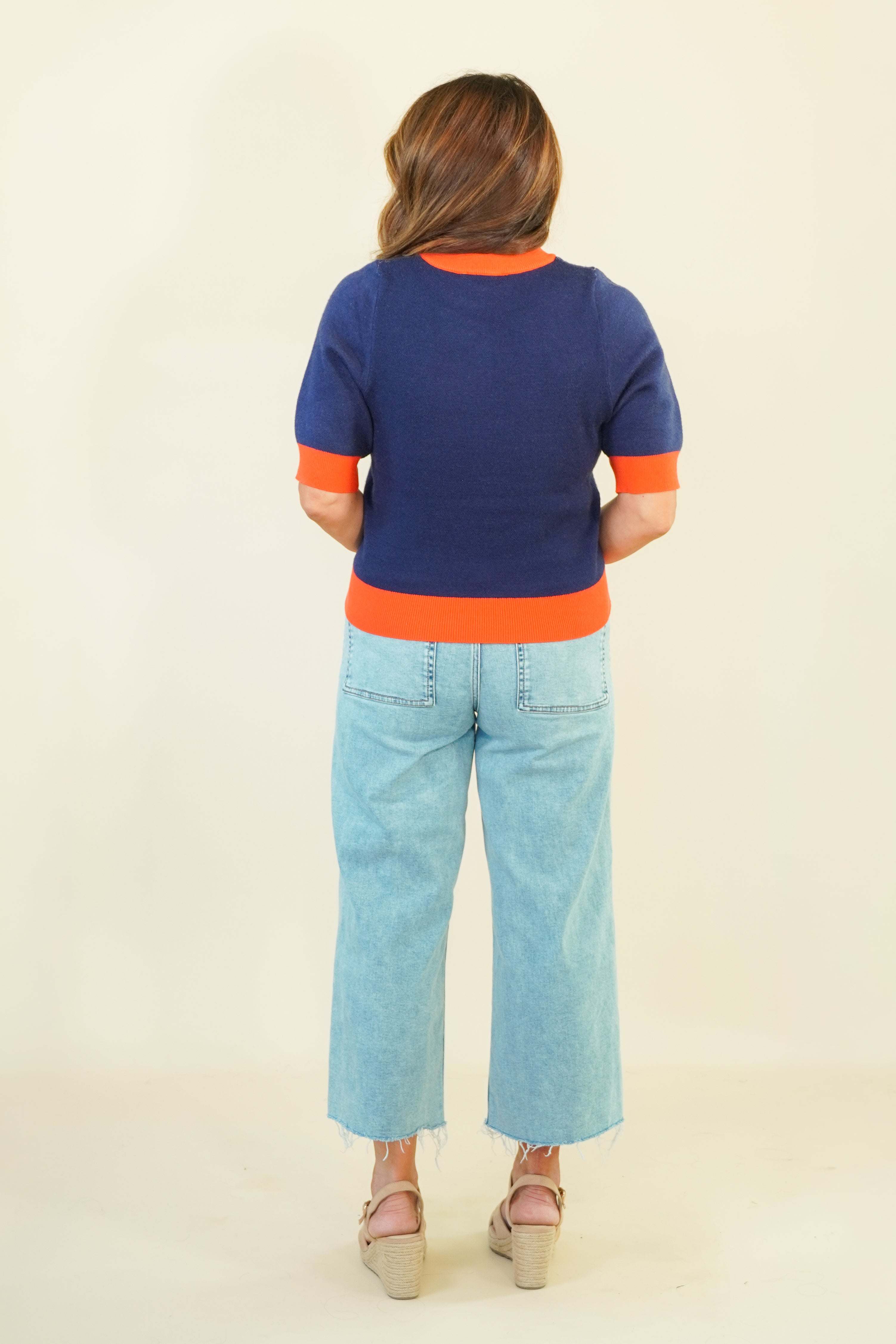 A woman viewed from behind wearing a short-sleeve knit sweater with a navy blue body and bright orange trim at the collar, cuffs, and hem, paired with light blue cropped jeans and beige wedge sandals, against a plain cream background.