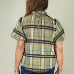 A woman viewed from the back wearing a short-sleeve plaid blouse in olive green, beige, and navy, paired with blue jeans, against a solid light gray background.