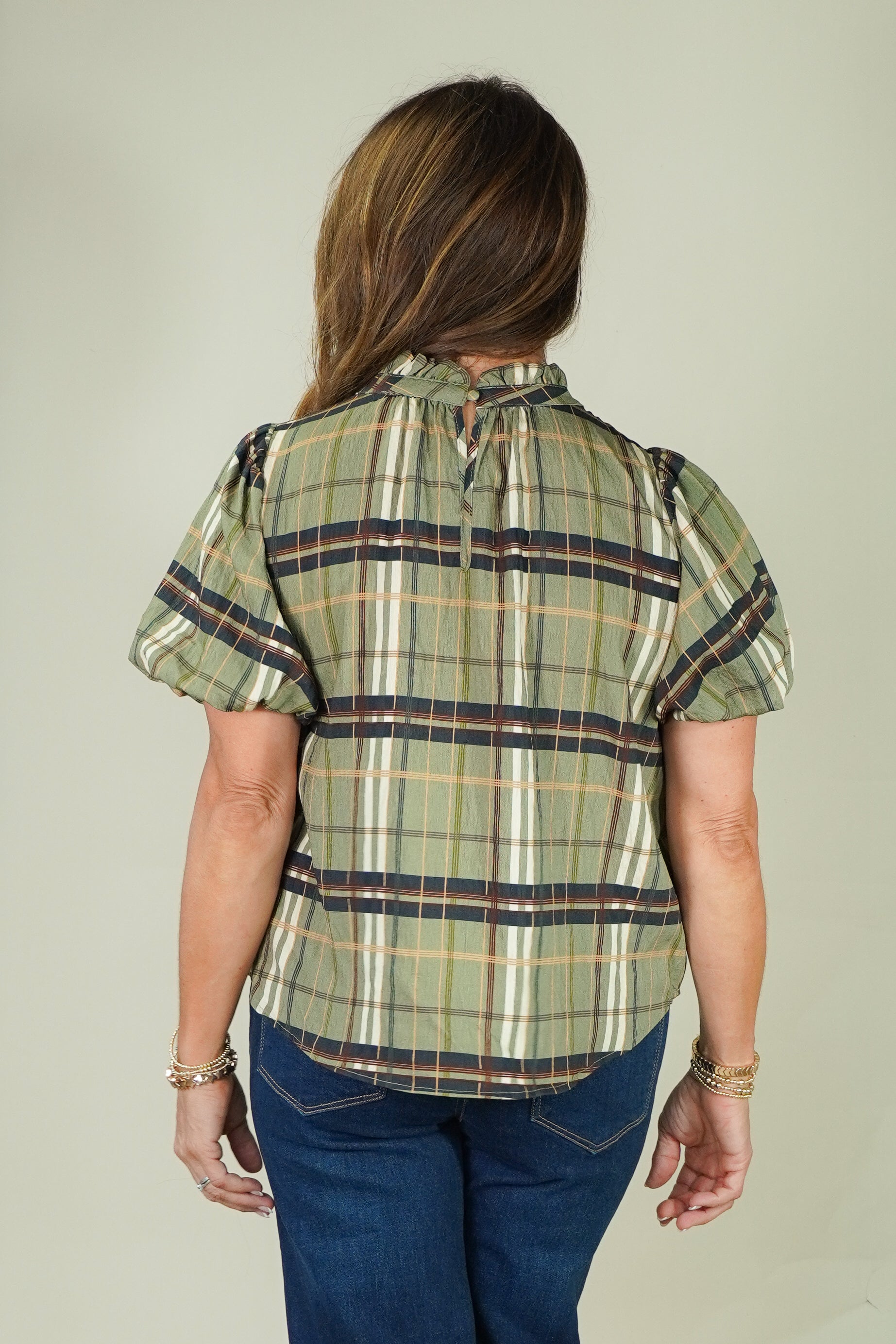 A woman viewed from the back wearing a short-sleeve plaid blouse in olive green, beige, and navy, paired with blue jeans, against a solid light gray background.