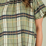 A woman with long brown hair smiling, wearing a short-sleeve plaid blouse in olive green, beige, and brown, paired with blue jeans, against a solid light beige background.