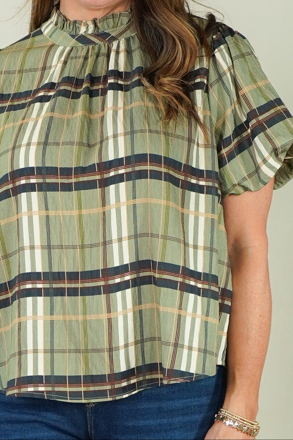 A woman with long brown hair smiling, wearing a short-sleeve plaid blouse in olive green, beige, and brown, paired with blue jeans, against a solid light beige background.