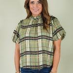 A woman wearing a short-sleeve plaid blouse with green, beige, and navy stripes, paired with blue jeans, standing against a plain light gray background.