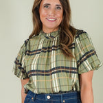 A woman with long brown hair smiling, wearing a short-sleeve plaid blouse in olive green, beige, and navy, paired with dark blue jeans, against a solid light gray background.