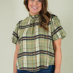 A woman wearing a short-sleeve plaid blouse in olive green, beige, and navy blue, paired with dark blue jeans, standing against a solid light gray background.