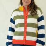 A woman modeling a colorful striped knit cardigan sweater with blue jeans and pink socks, standing against a plain white studio background.