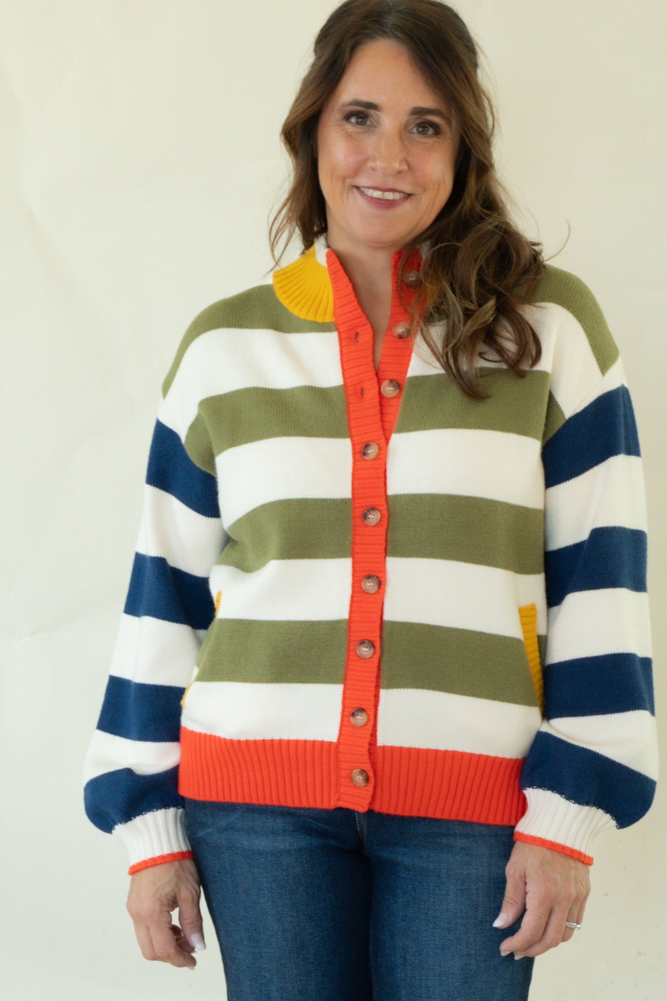 A woman modeling a colorful striped knit cardigan sweater with blue jeans and pink socks, standing against a plain white studio background.