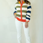 A woman modeling a colorful striped cardigan sweater with white pants and brown woven loafers, standing against a plain off-white studio background.
