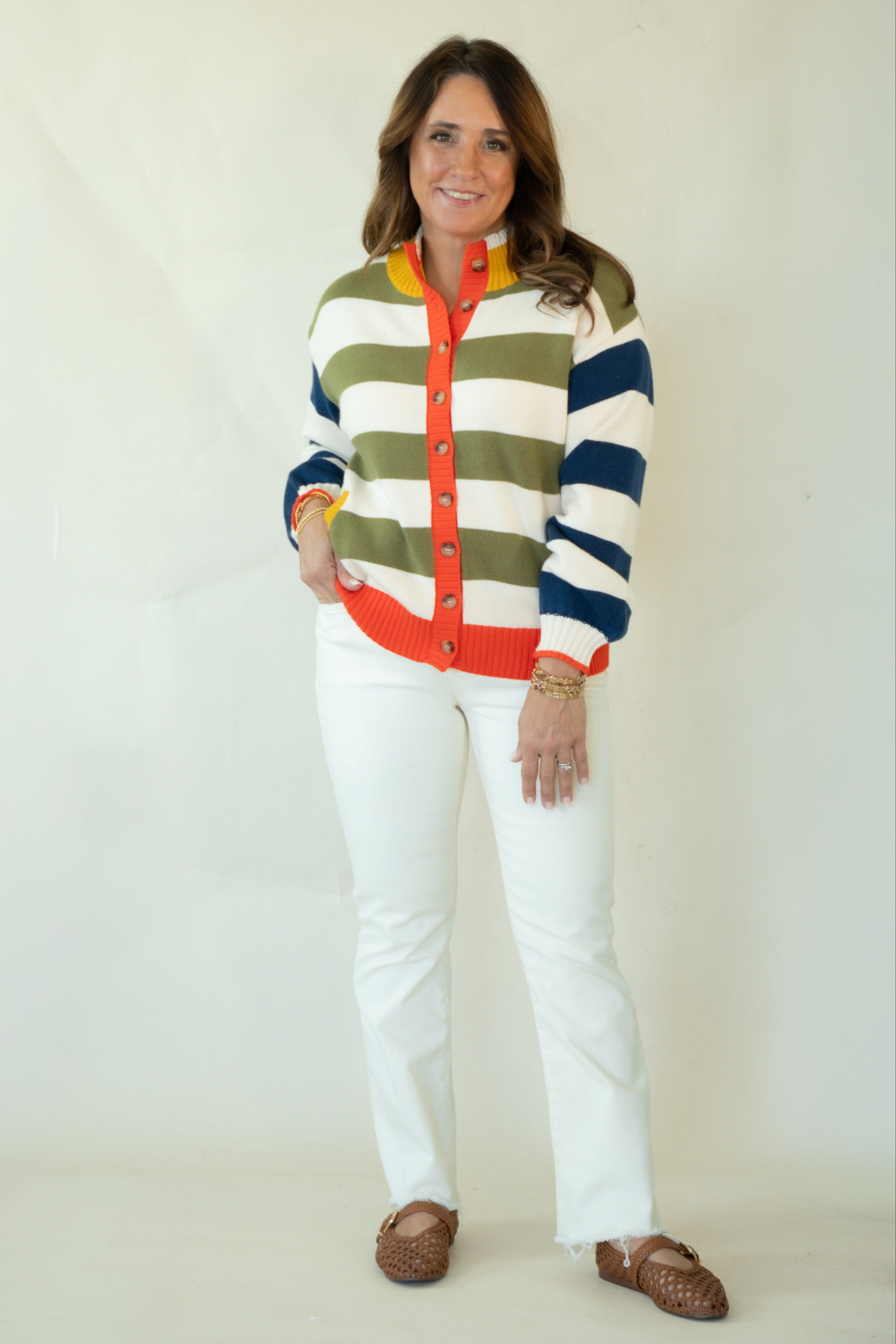 A woman modeling a colorful striped cardigan sweater with white pants and brown woven loafers, standing against a plain off-white studio background.