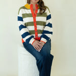 A woman sits barefoot on a white textured cylindrical stool, wearing a colorful striped cardigan with navy, olive green, white, and yellow horizontal stripes, a red button placket, and blue jeans against a plain off-white studio background.