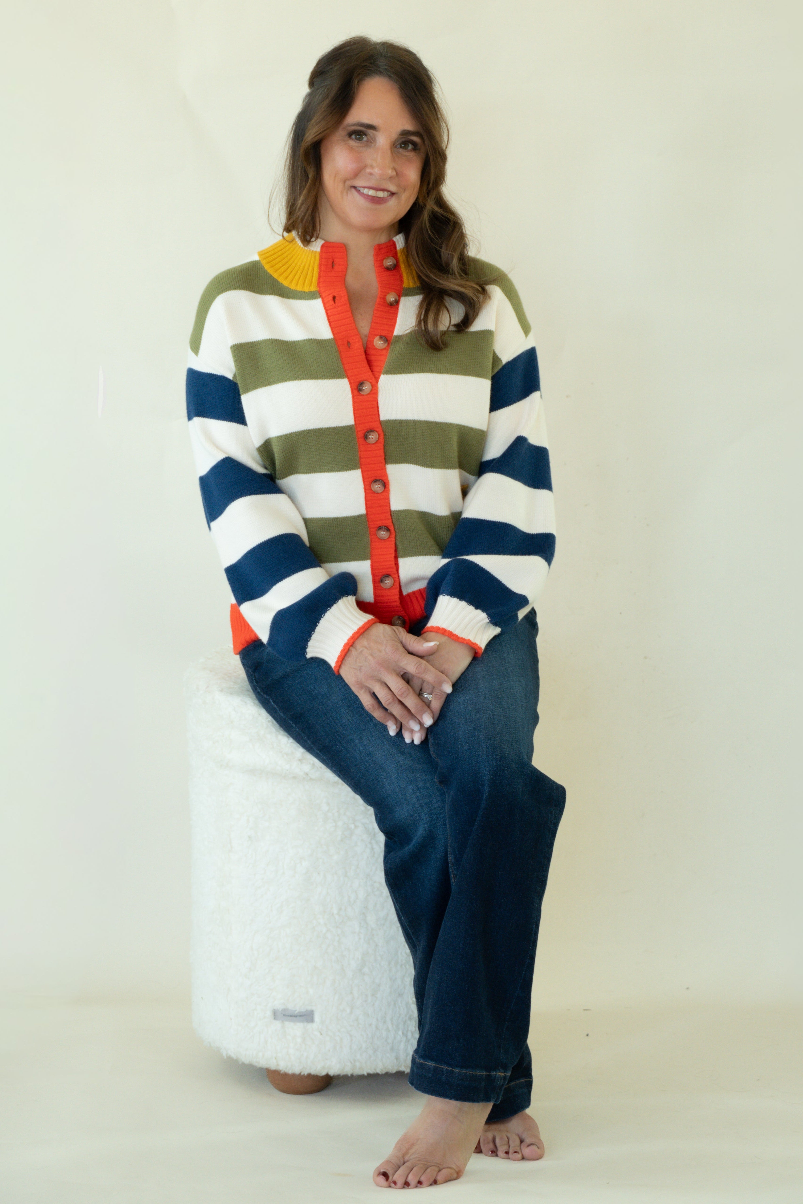 A woman sits barefoot on a white textured cylindrical stool, wearing a colorful striped cardigan with navy, olive green, white, and yellow horizontal stripes, a red button placket, and blue jeans against a plain off-white studio background.