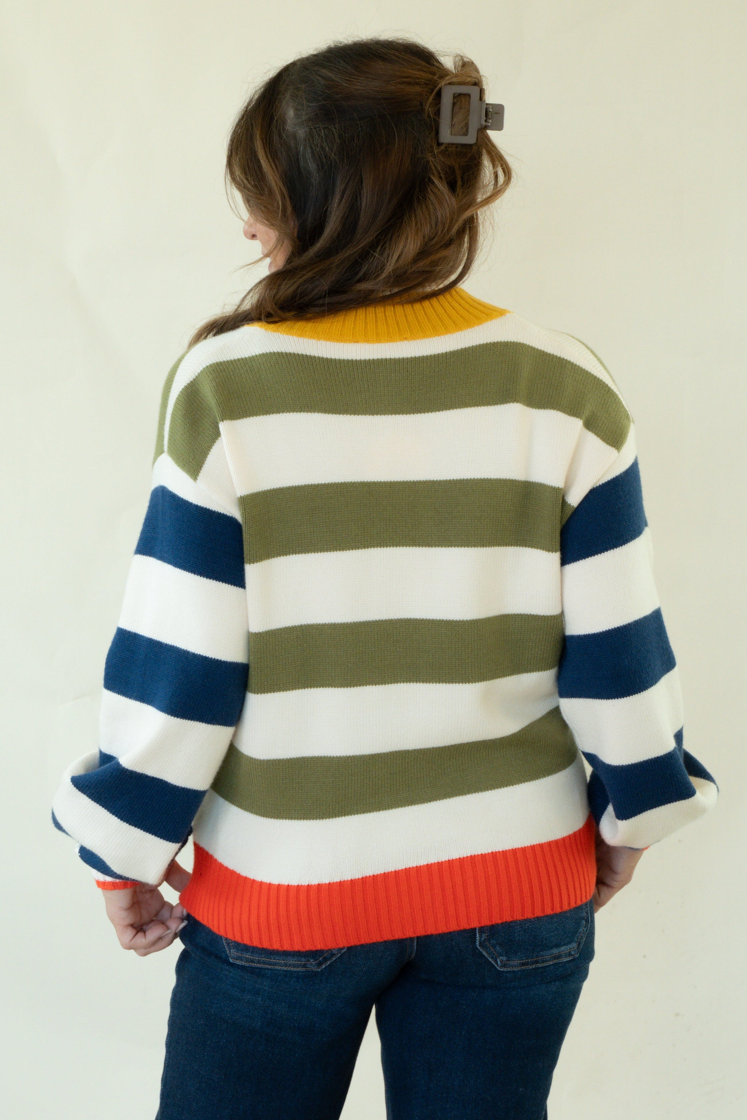 A woman viewed from behind wearing a striped knit sweater with navy blue, olive green, white, and orange horizontal bands, paired with dark blue jeans against a plain off-white studio background.