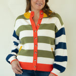 A woman stands barefoot against a plain off-white studio background wearing a striped knit cardigan with blue, olive green, white, and orange horizontal bands, paired with dark blue jeans.